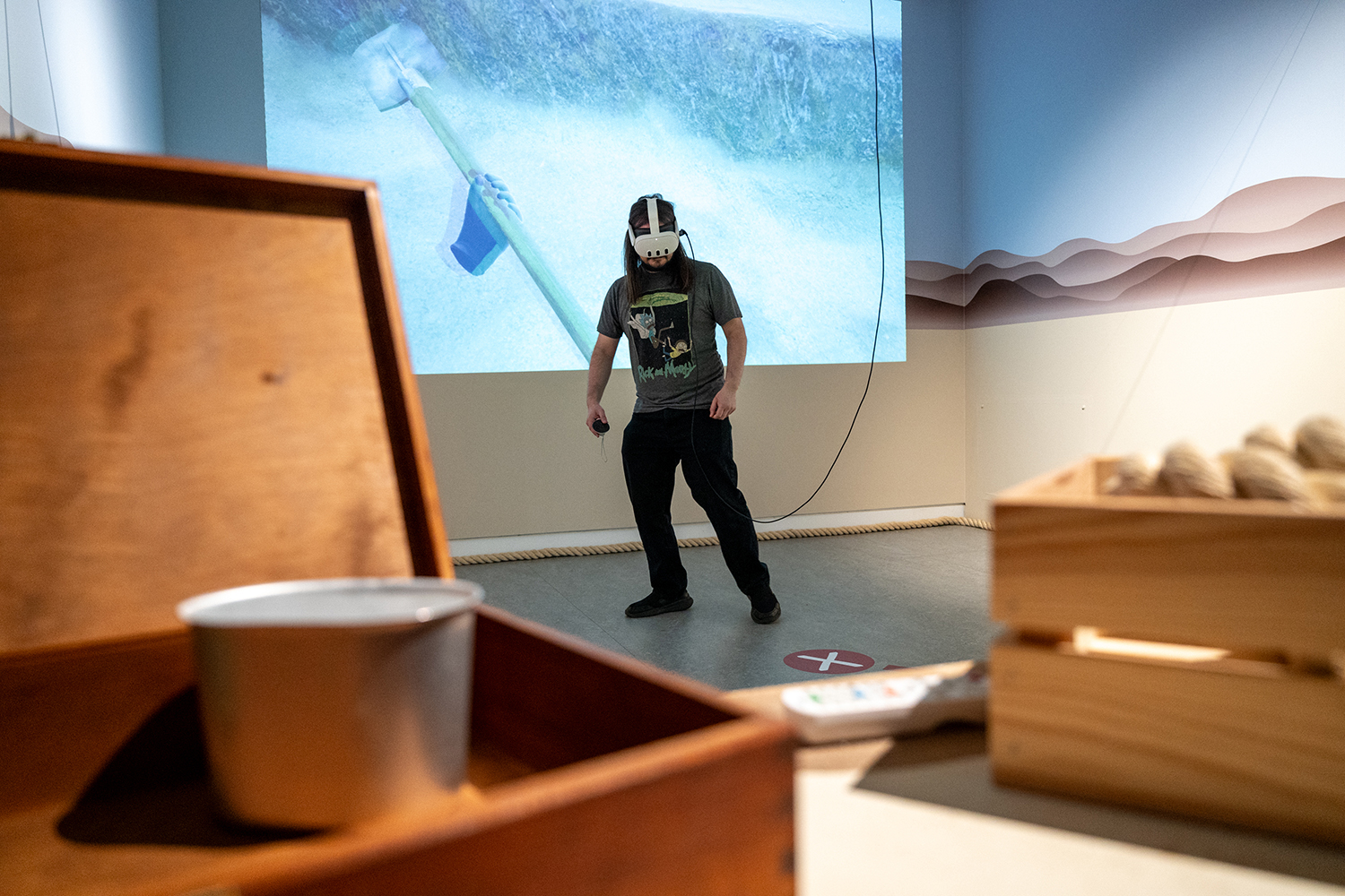 A man wears a VR headset with a projection behind him of an archaeological dig site in a gallery